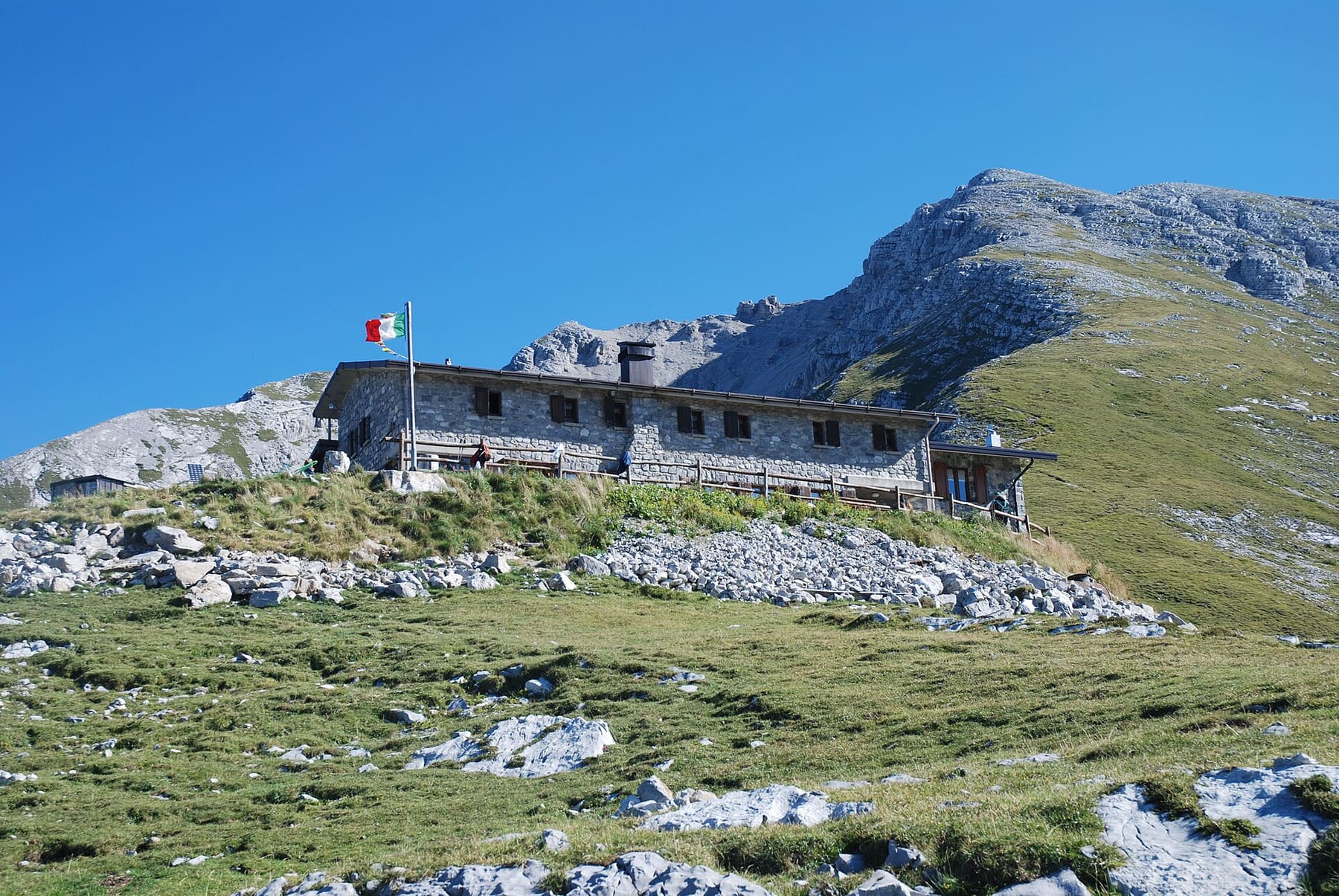 Il rifugio Capanna 2000 in pietra con la bandiera italiana sui pascoli dell'Alpe Arera, sotto le rocce del pizzo Arera