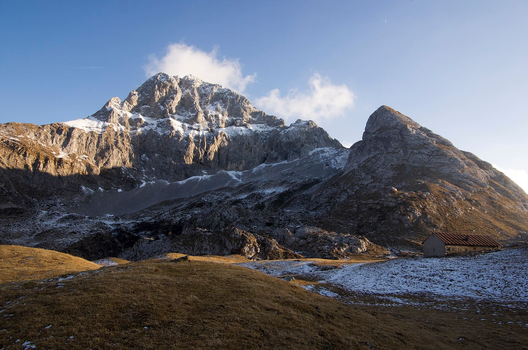 Le pareti rocciose del Pizzo Arera e della Corna Piana spolverate di neve nella luce del tardo pomeriggio, viste dal Sentiero dei Fiori, con una baita tra i pascoli