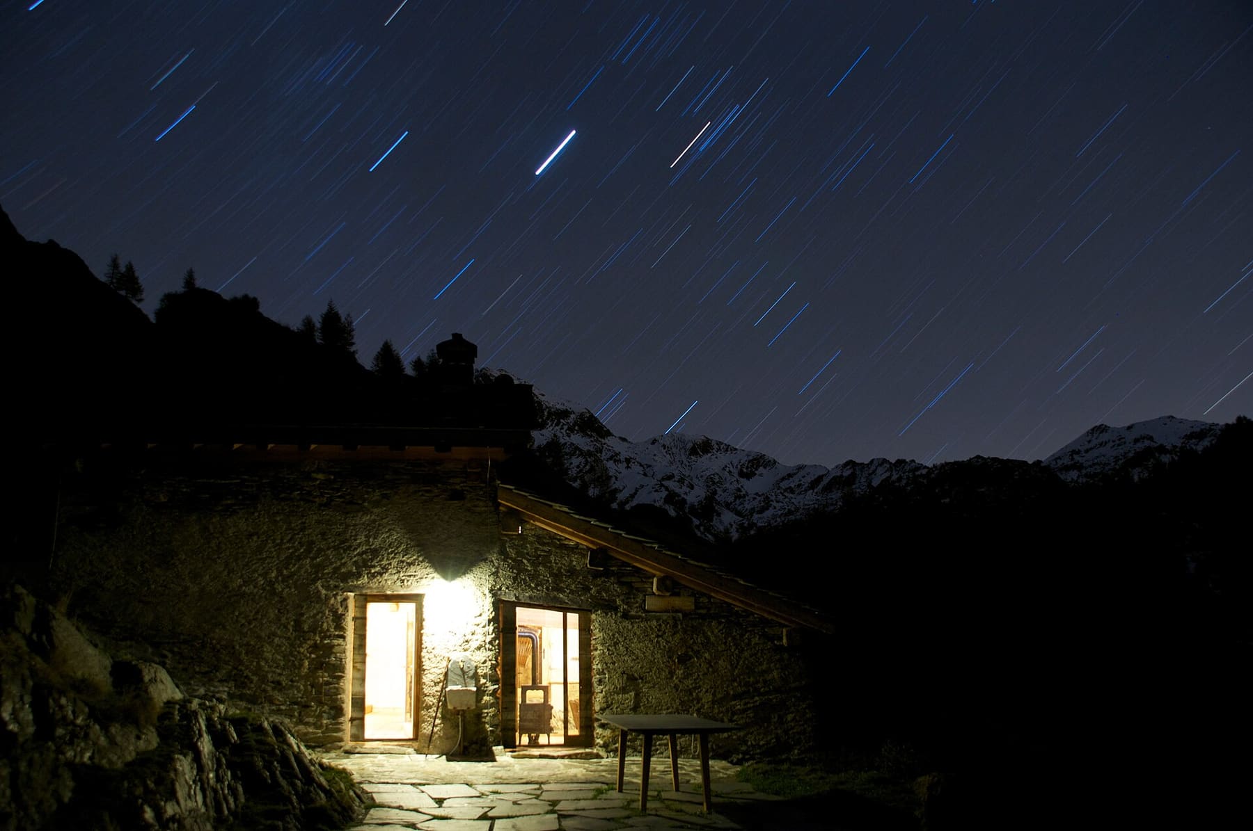 Baita in pietra illuminata di notte sotto un cielo stellato, con le cime innevate delle Orobie in Val Brembana